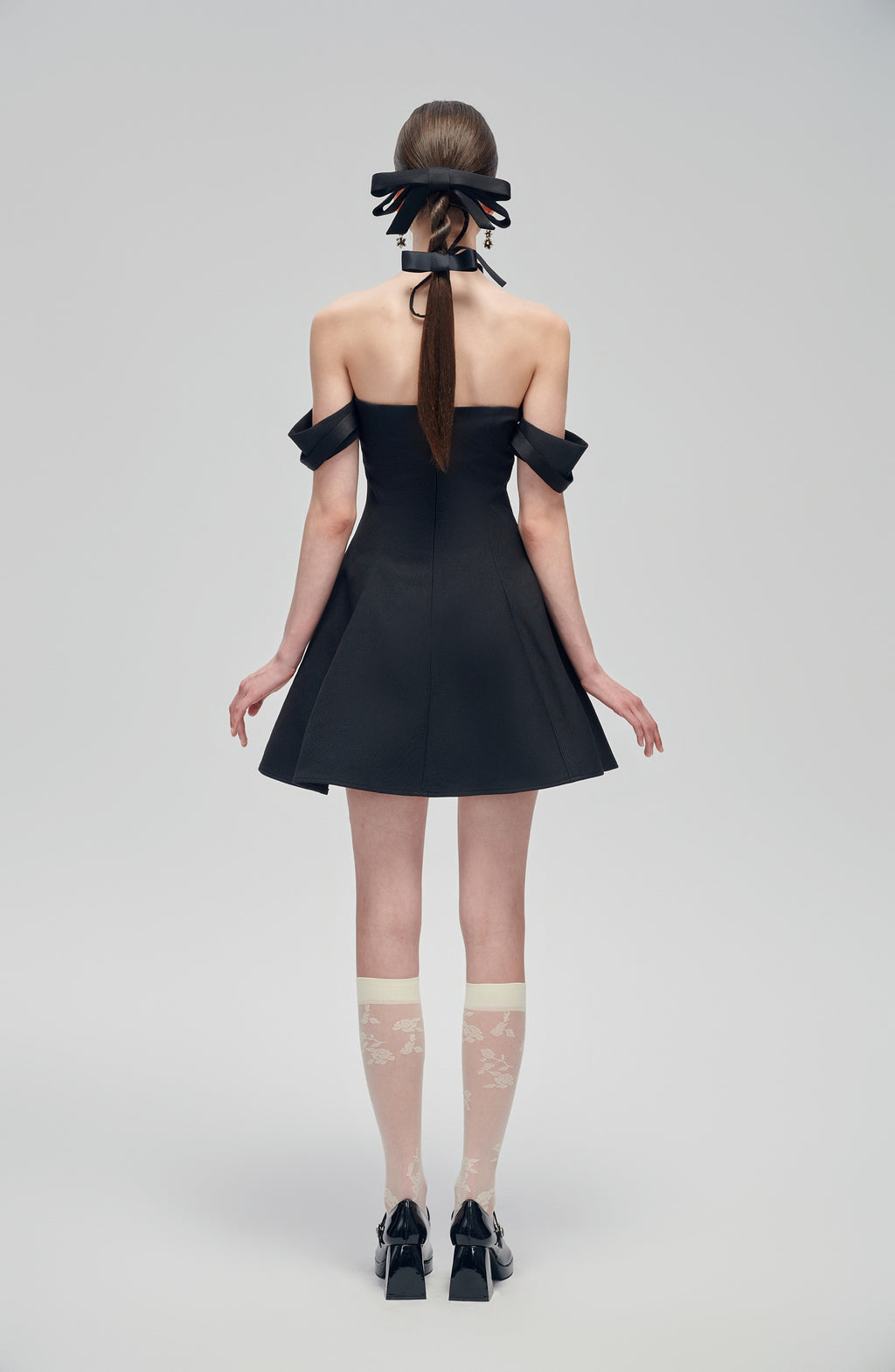 Back view of Nero Ritratto Off-Shoulder Dress featuring double off-shoulder straps and A-line silhouette in black. Styled with bow heels and sheer socks.