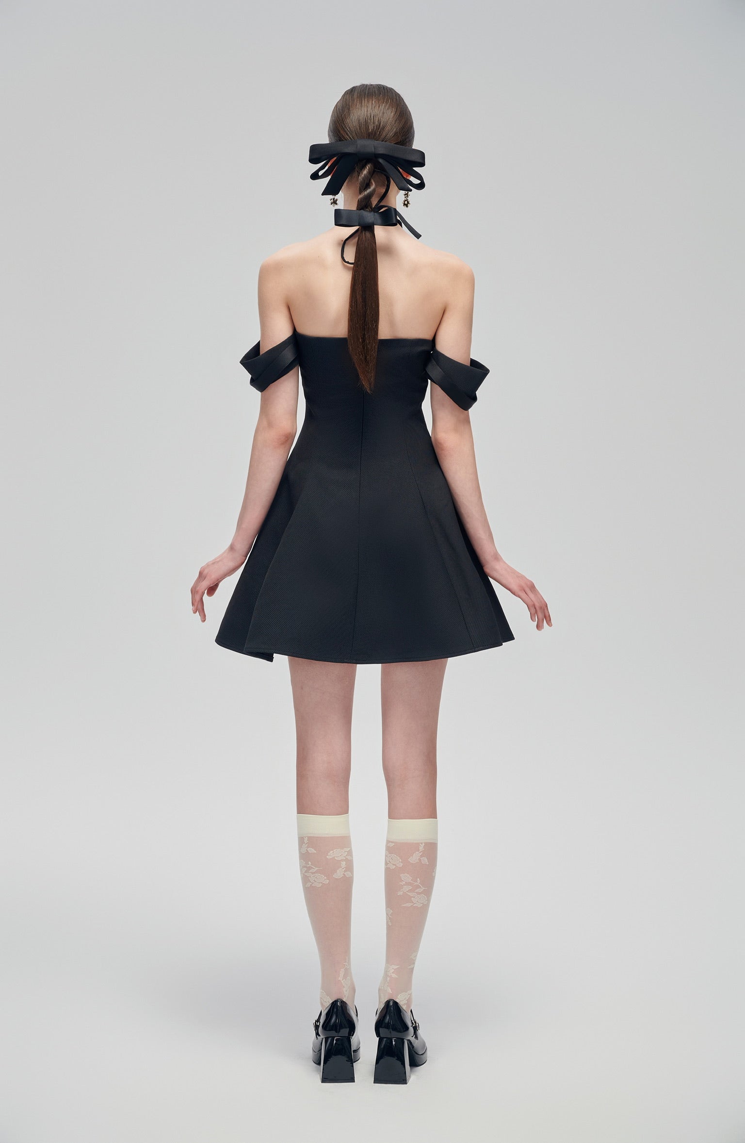 Back view of Nero Ritratto Off-Shoulder Dress featuring double off-shoulder straps and A-line silhouette in black. Styled with bow heels and sheer socks.